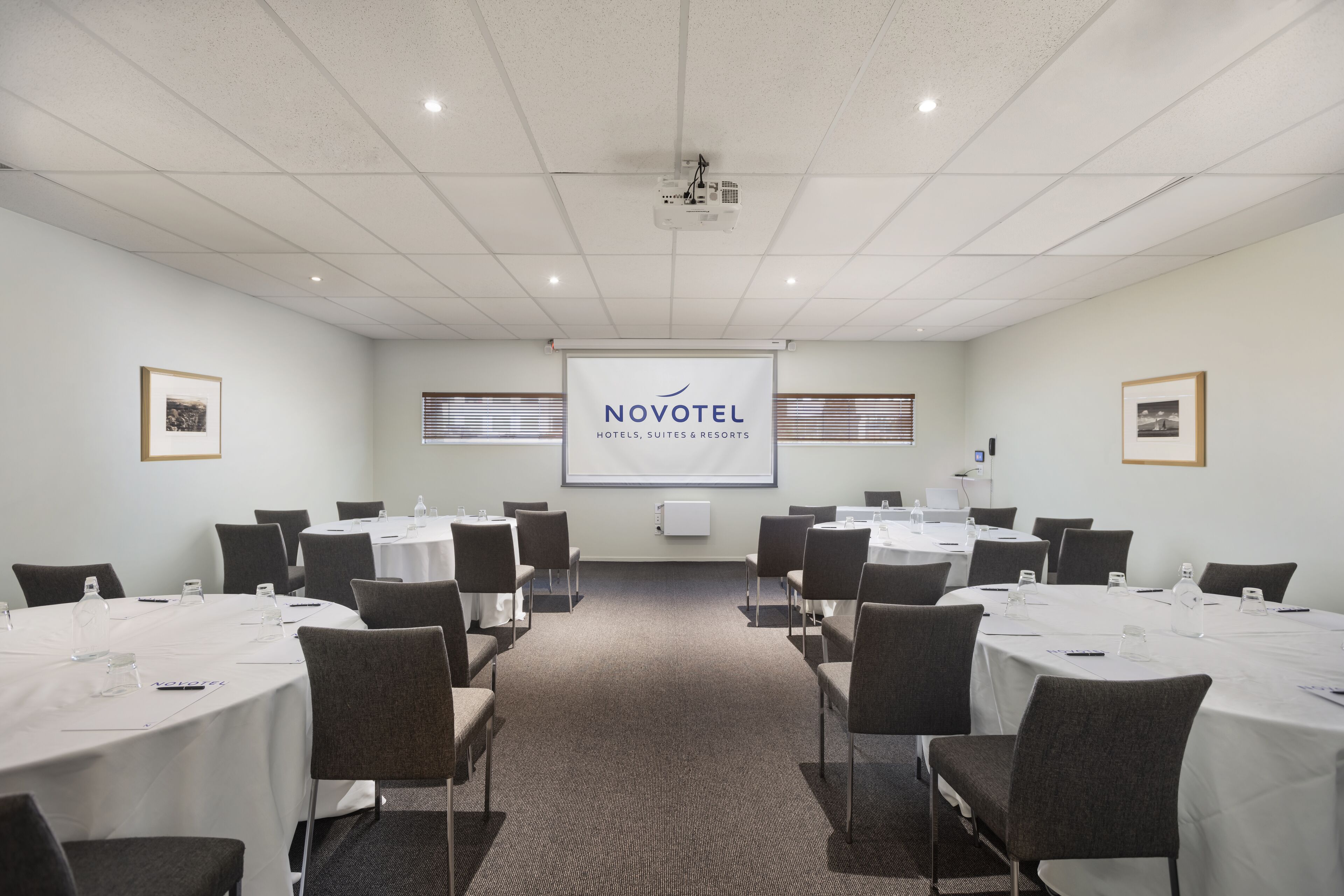 A conference room with multiple tables and chairs, a projector screen, and framed pictures on the walls.