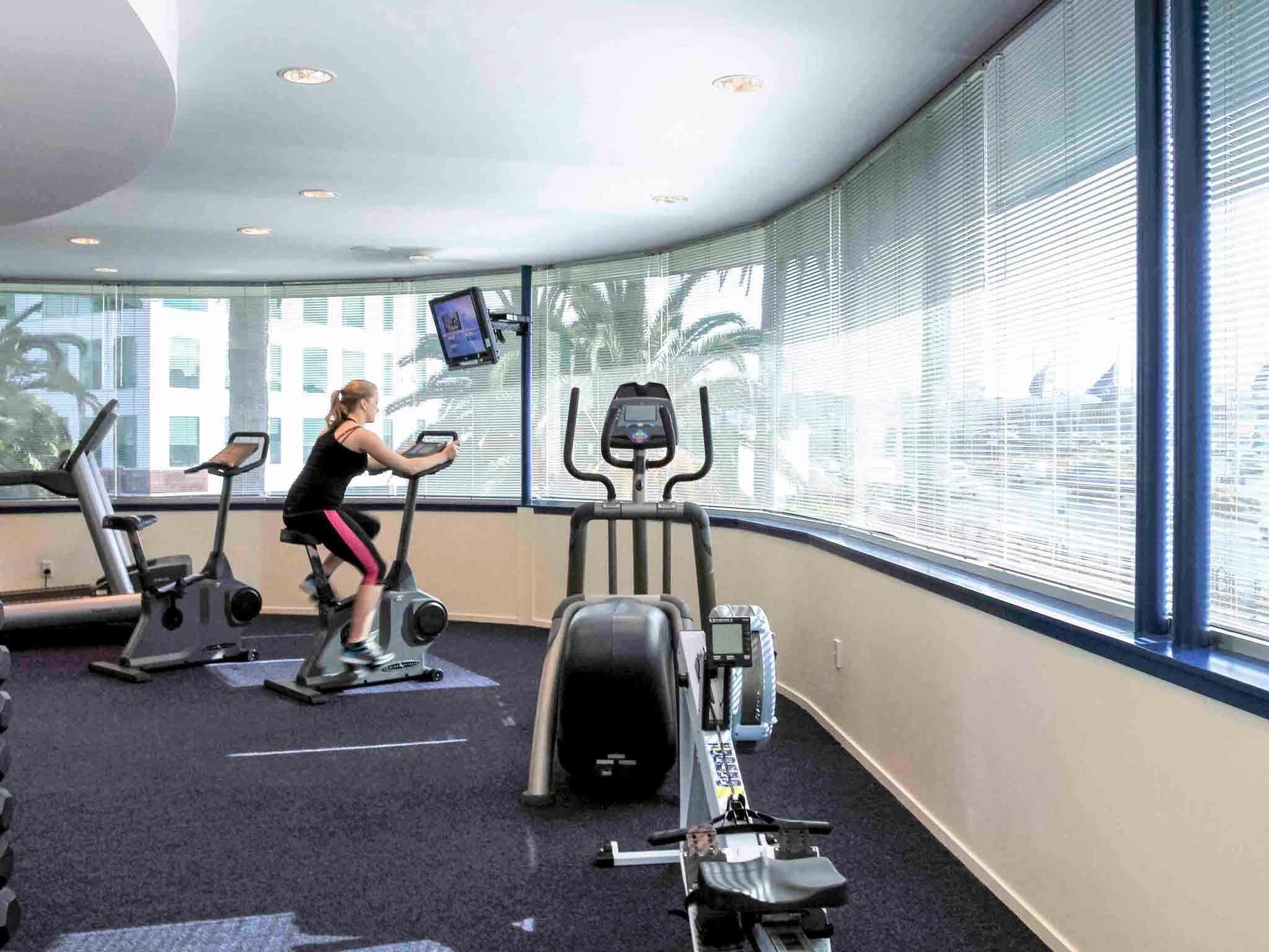 Ein Fitnessraum mit Cardiogeräten, einer Gewichtshalterung und einem großen Fenster mit Jalousien.