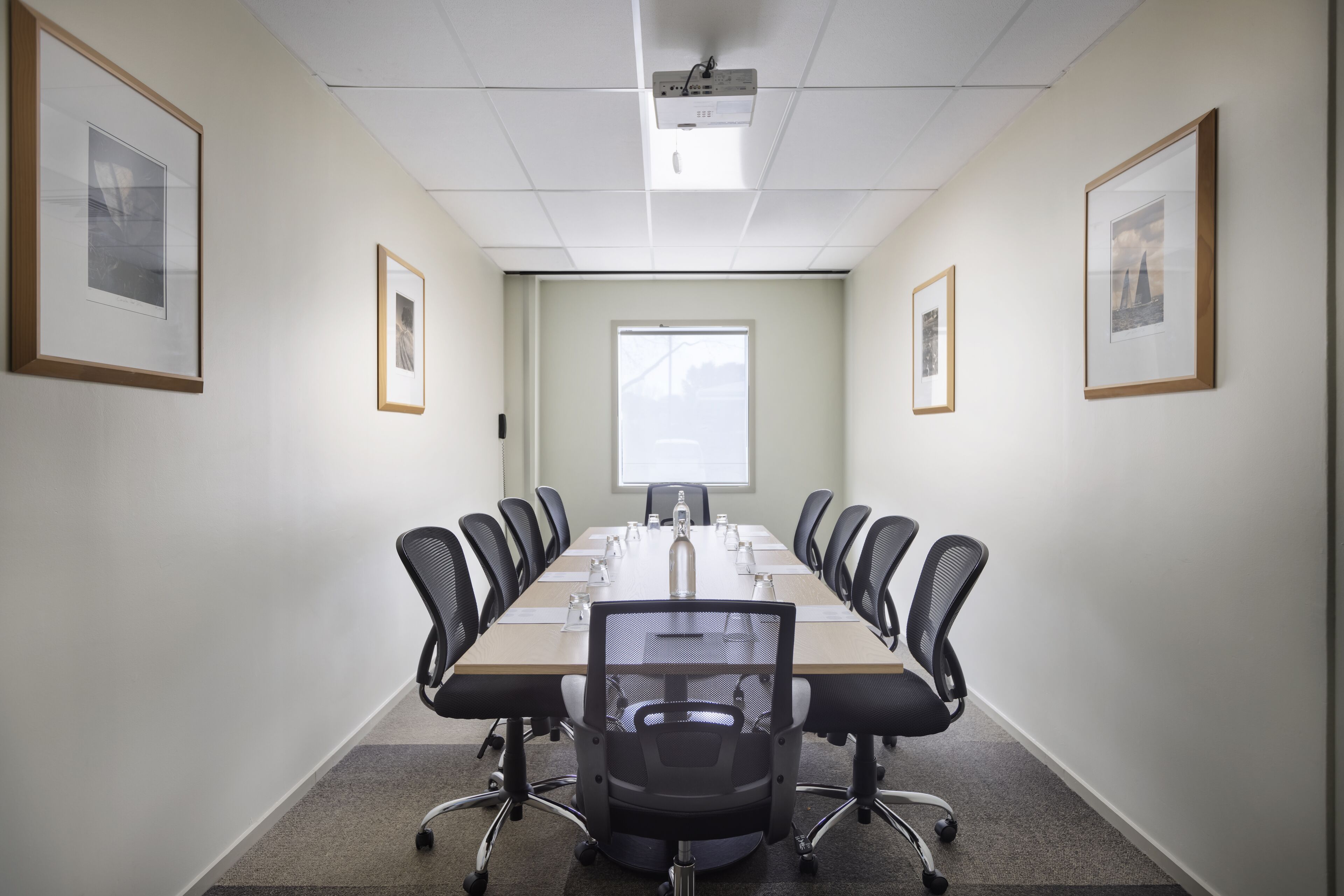 A conference room with a long table, black chairs, framed pictures on the walls, and a projector on the ceiling.