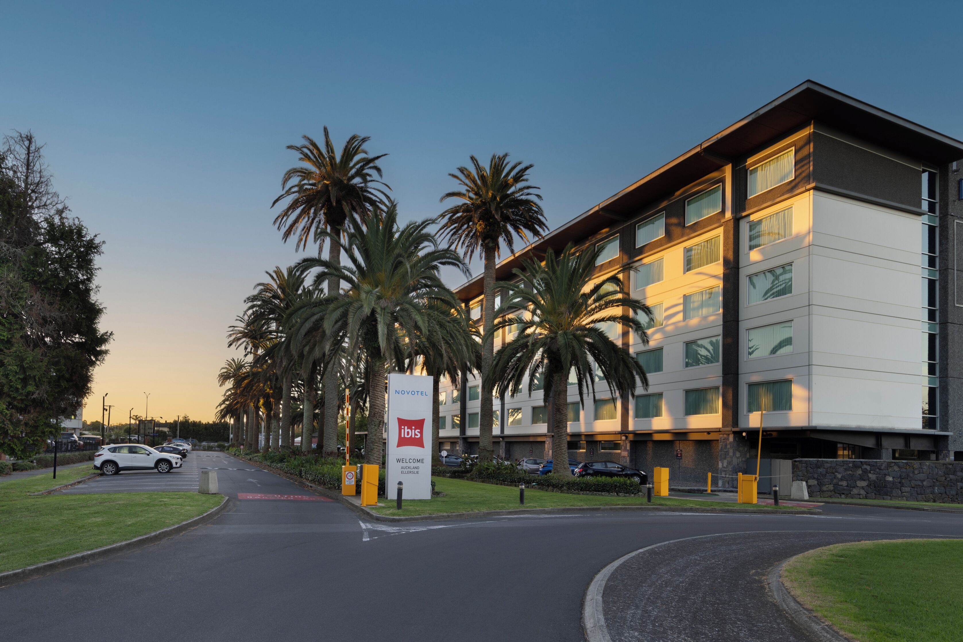 A modern hotel with a welcoming sign, palm trees, and a parking area.
