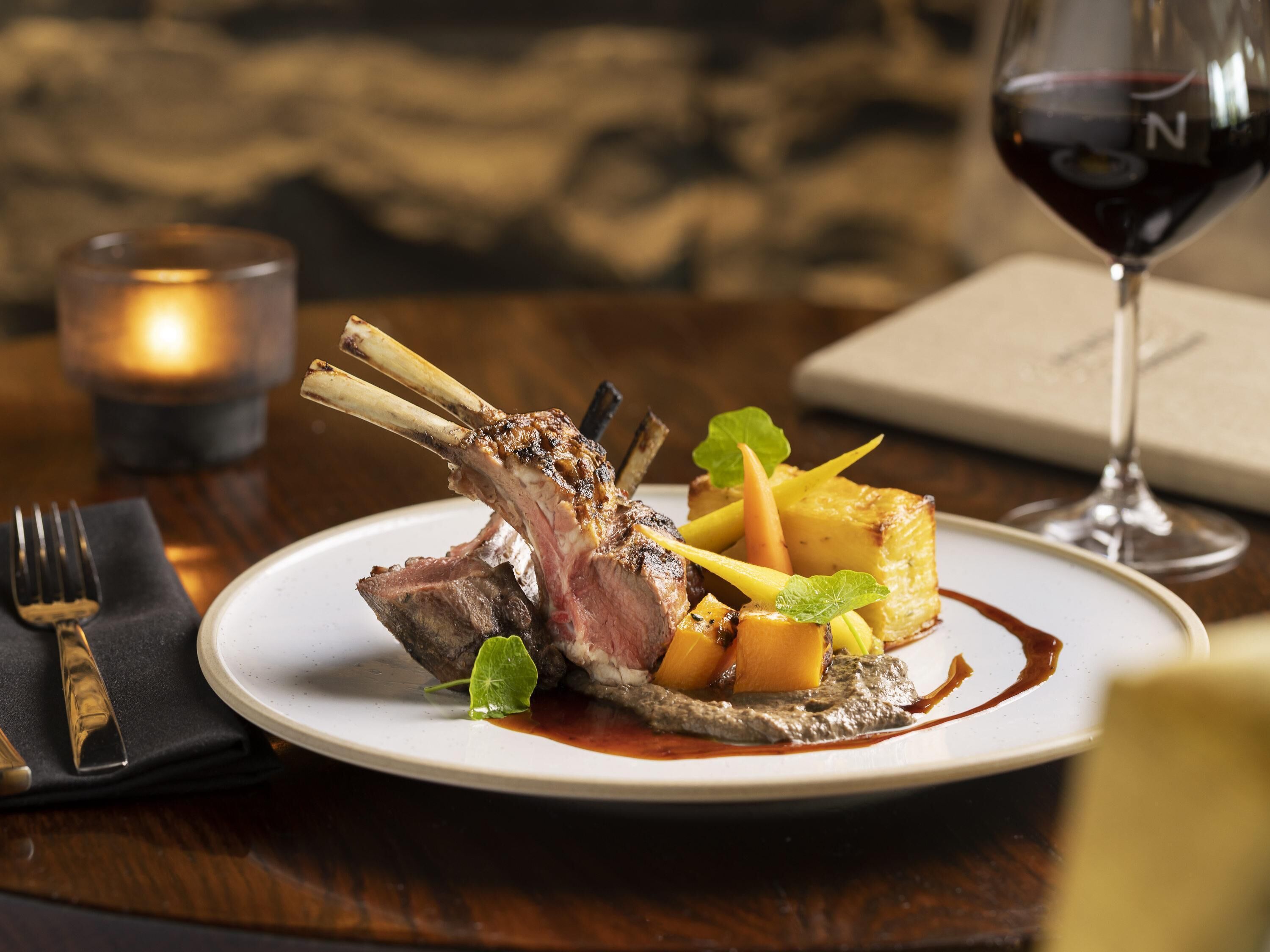 A plate of food with a lamb shank, vegetables, and sauce, accompanied by a glass of red wine and a candle.
