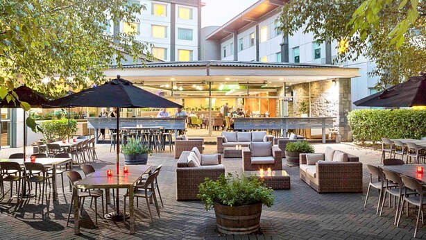 Een terras buiten met tafels en stoelen, omgeven door potplanten en parasols.