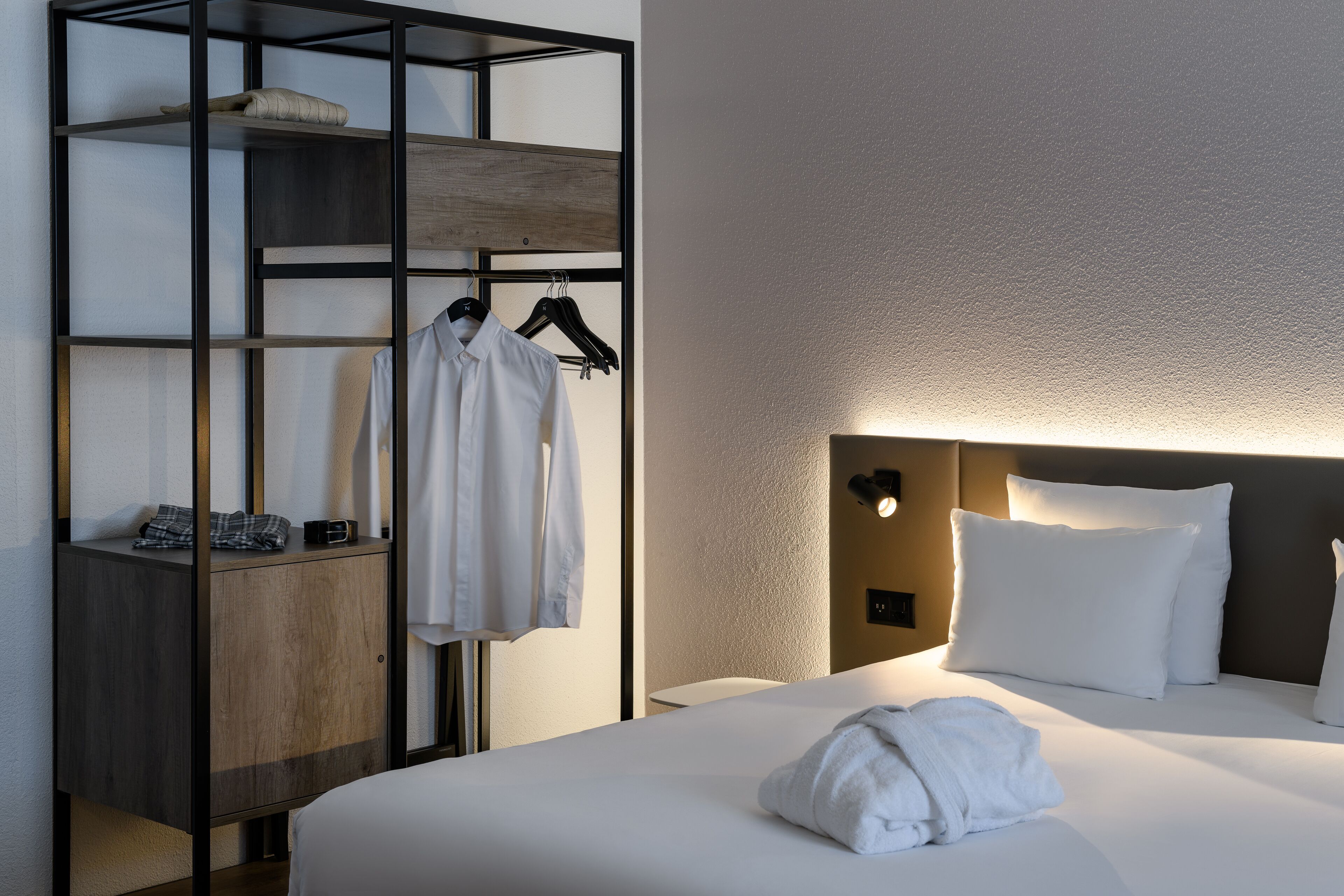Una habitación de hotel con una cama, un armario de madera y una percha con ropa.