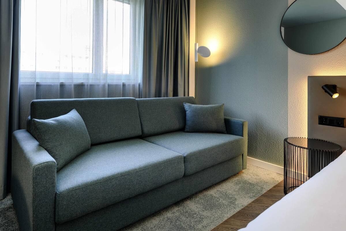 Moderni hotellihuone, jossa on harmaa sohva, pyöreä peili ja pieni sivupöytä.