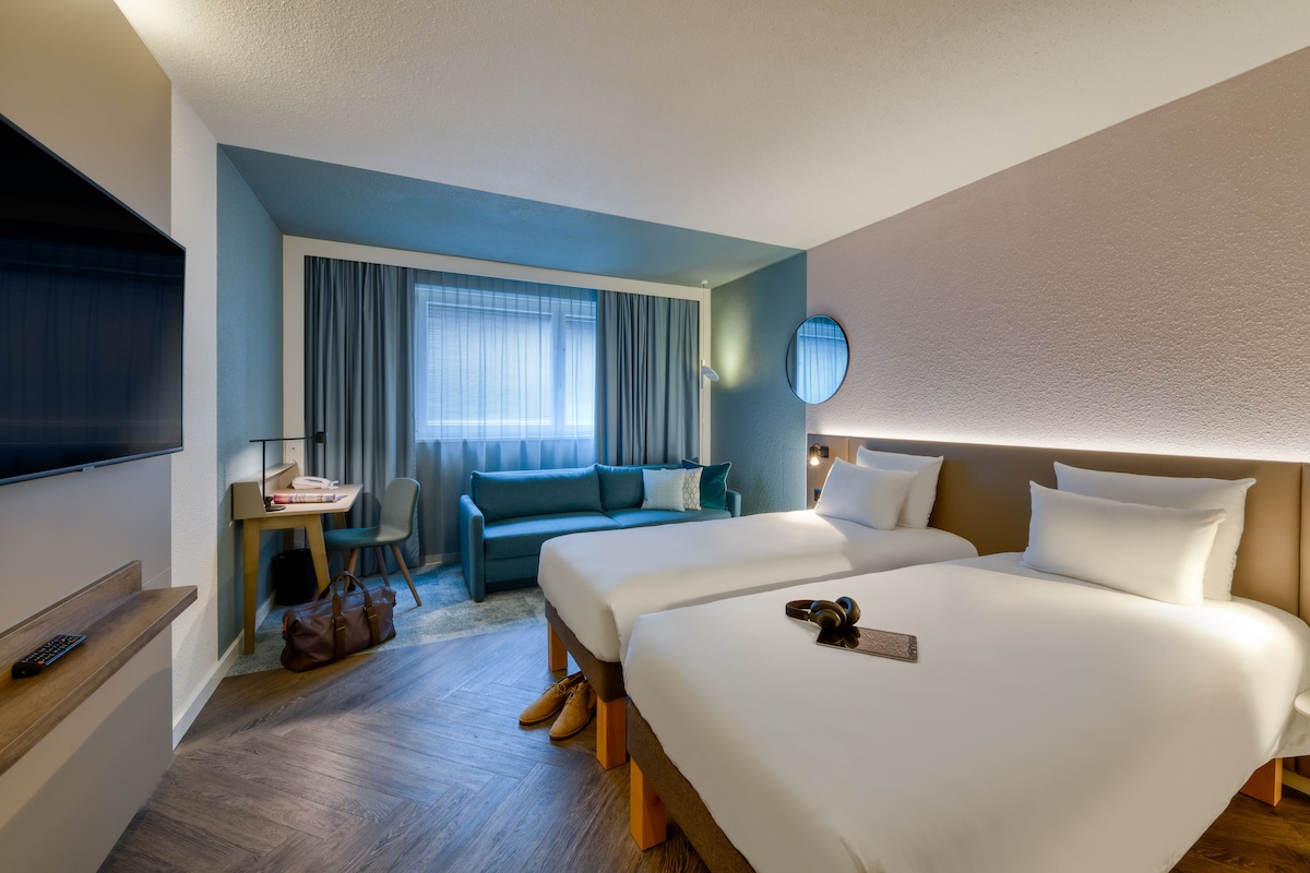 Hotellihuone, jossa on kaksi sänkyä, televisio, työpöytä tuolilla ja sohva.
