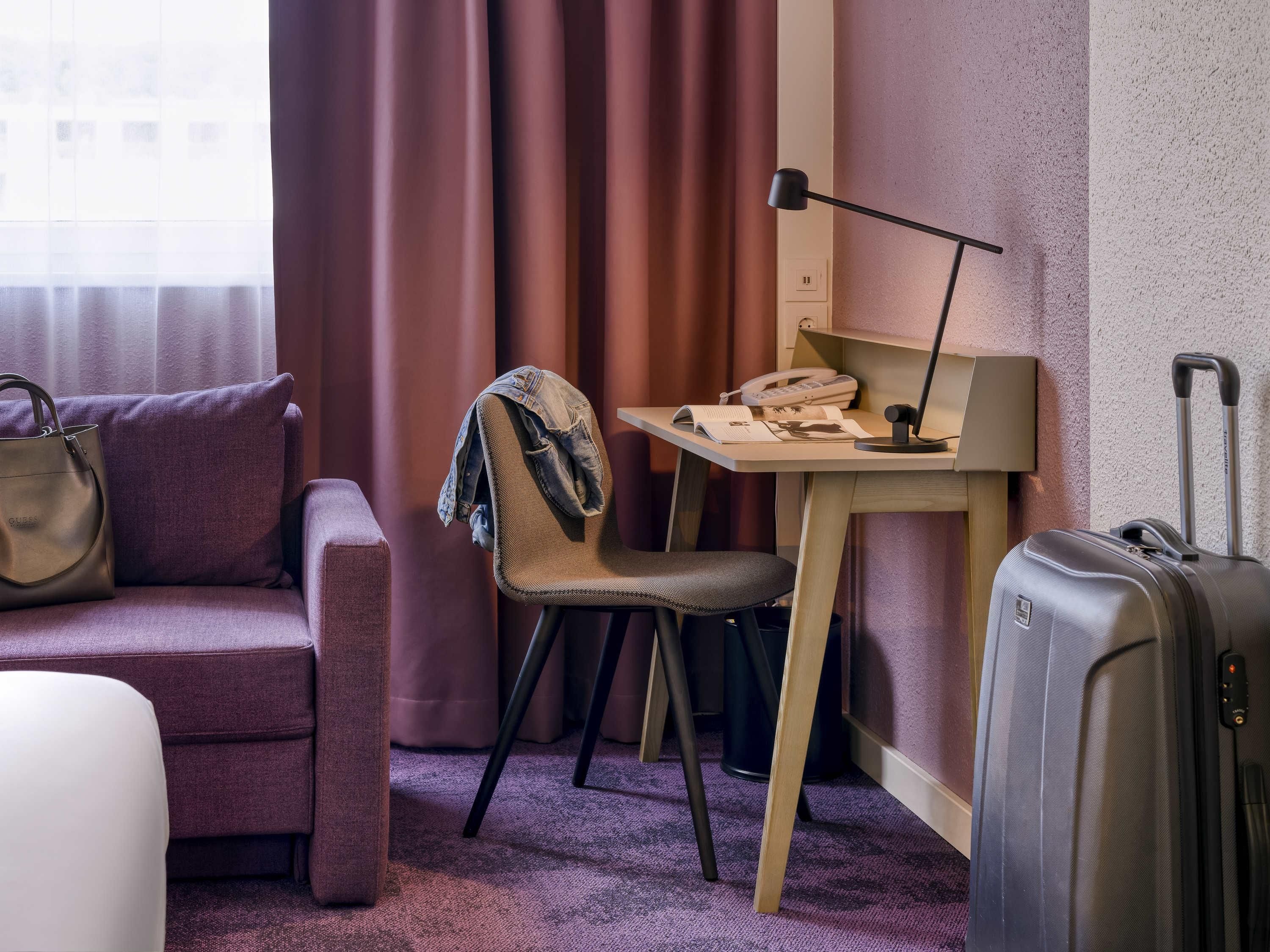 Quarto de hotel com sofá, escrivaninha com luminária, cadeira, mala e janela com cortinas.