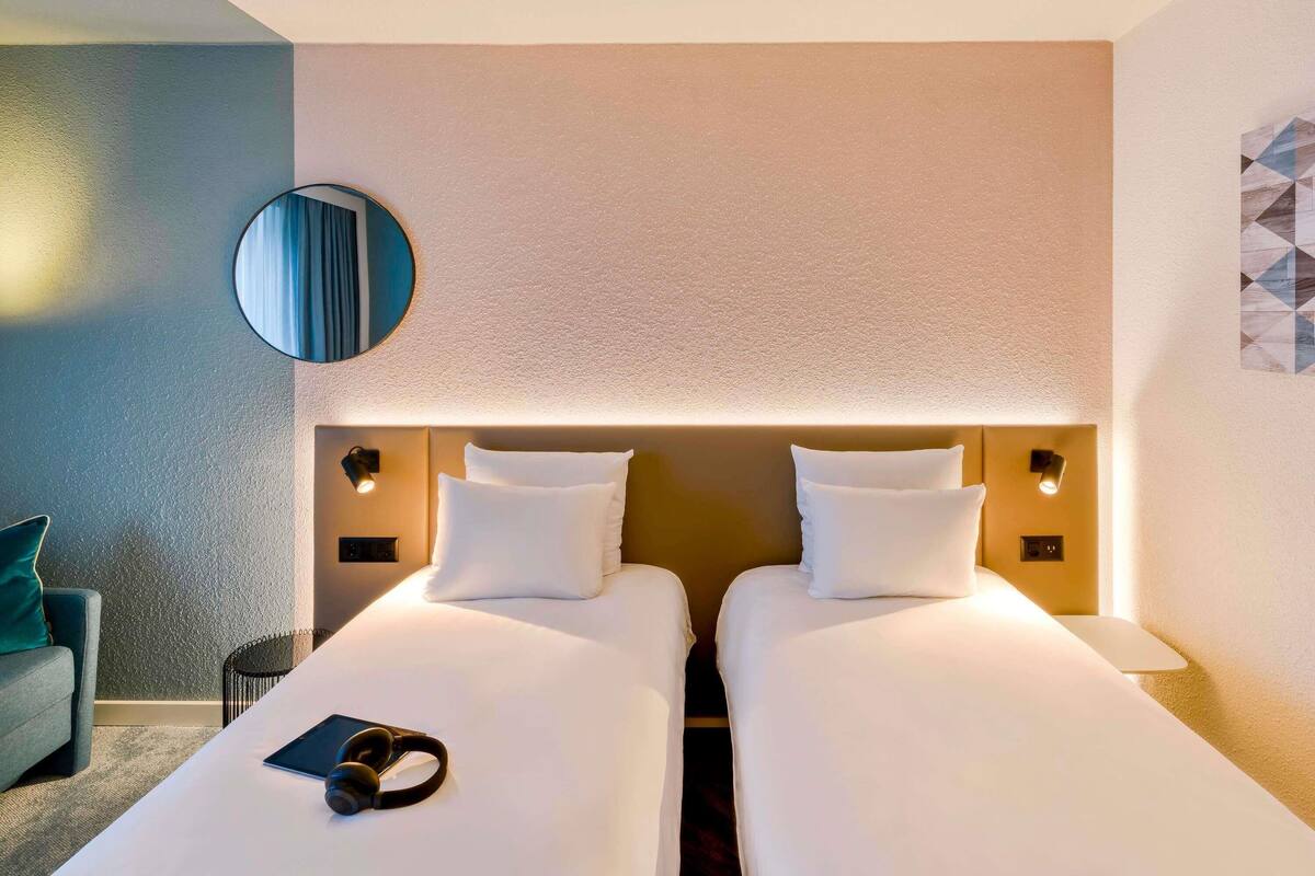 Hotellihuone, jossa on kaksi sänkyä, pyöreä peili, yöpöytävalaisimet, pieni pöytä ja tuoli.