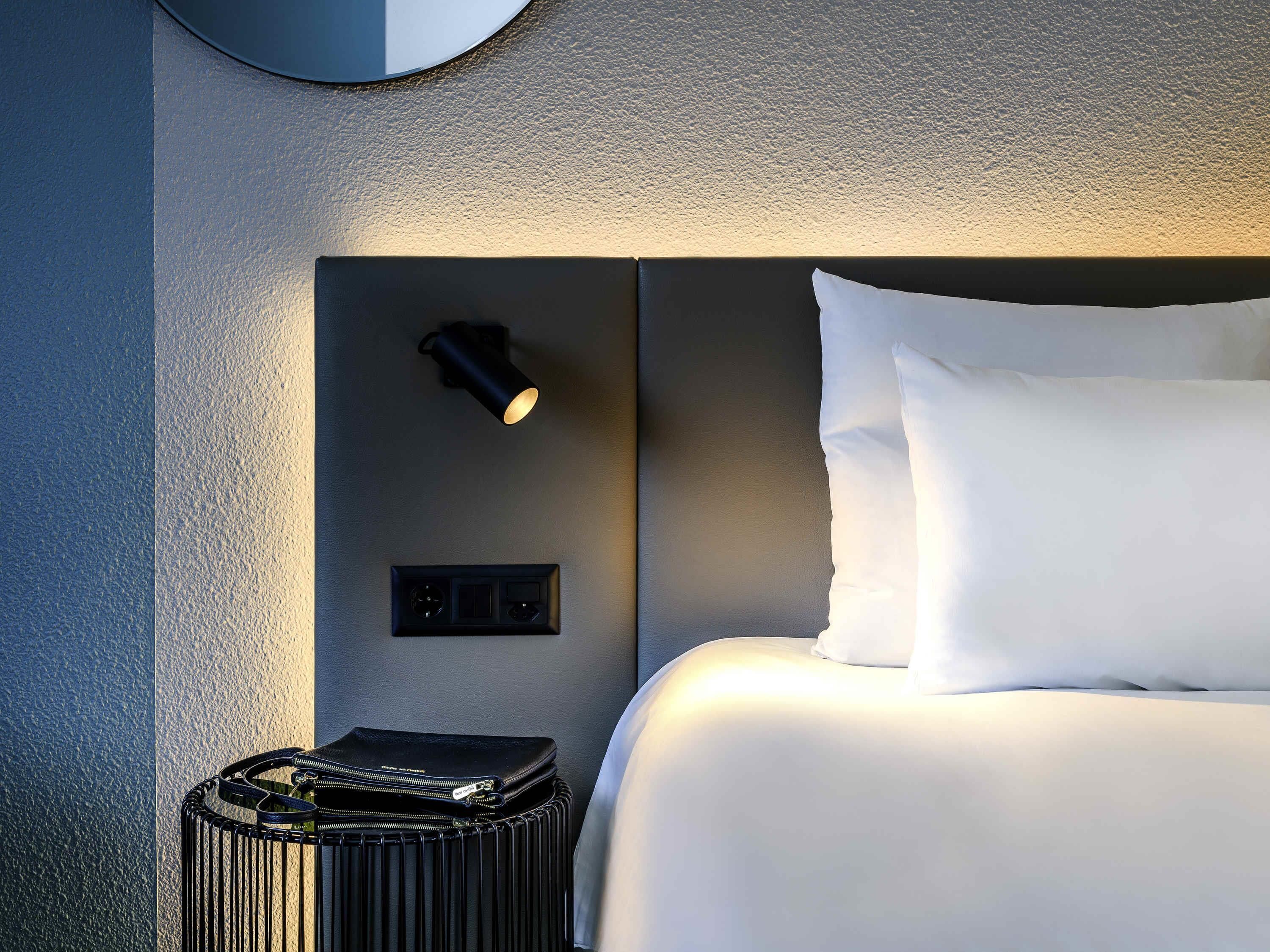 Un lit bien fait, avec du linge de lit blanc, une table de chevet noire avec une lampe, et un petit sac noir posé à côté.