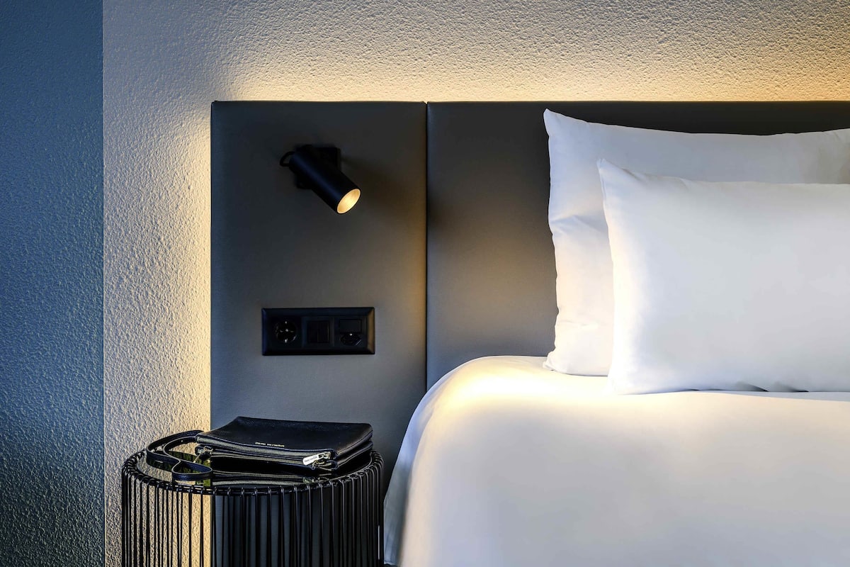 Un lit bien fait, avec du linge de lit blanc, une table de chevet noire avec une lampe, et un petit sac noir posé à côté.