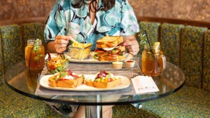 Een persoon zit aan een ronde glazen tafel met een maaltijd bestaande uit sandwiches, friet en een salade, samen met twee drankjes in weckpotten.