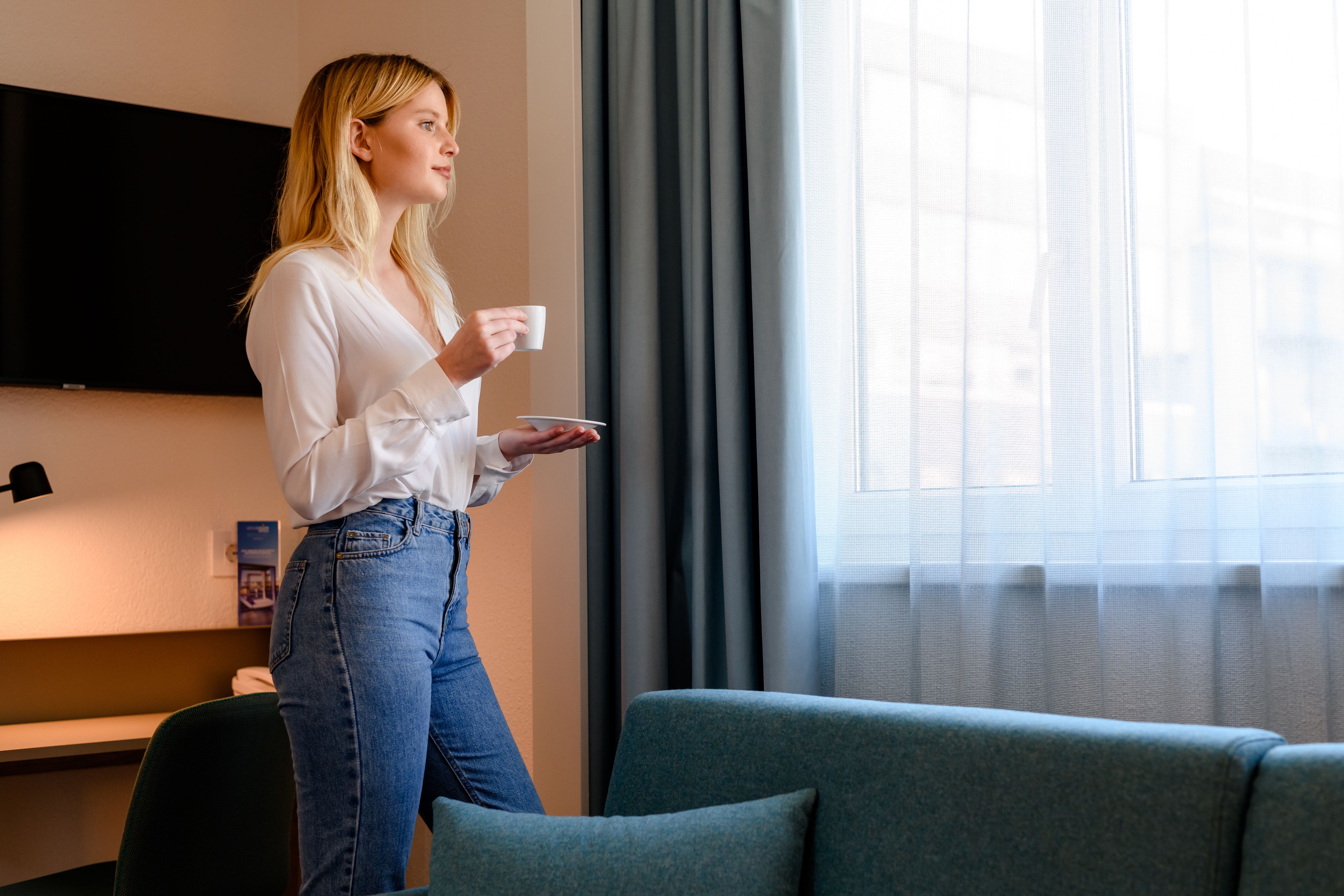 Una persona de pie en una habitación con un televisor de pantalla plana en la pared, una ventana con cortinas y un sofá verde.
