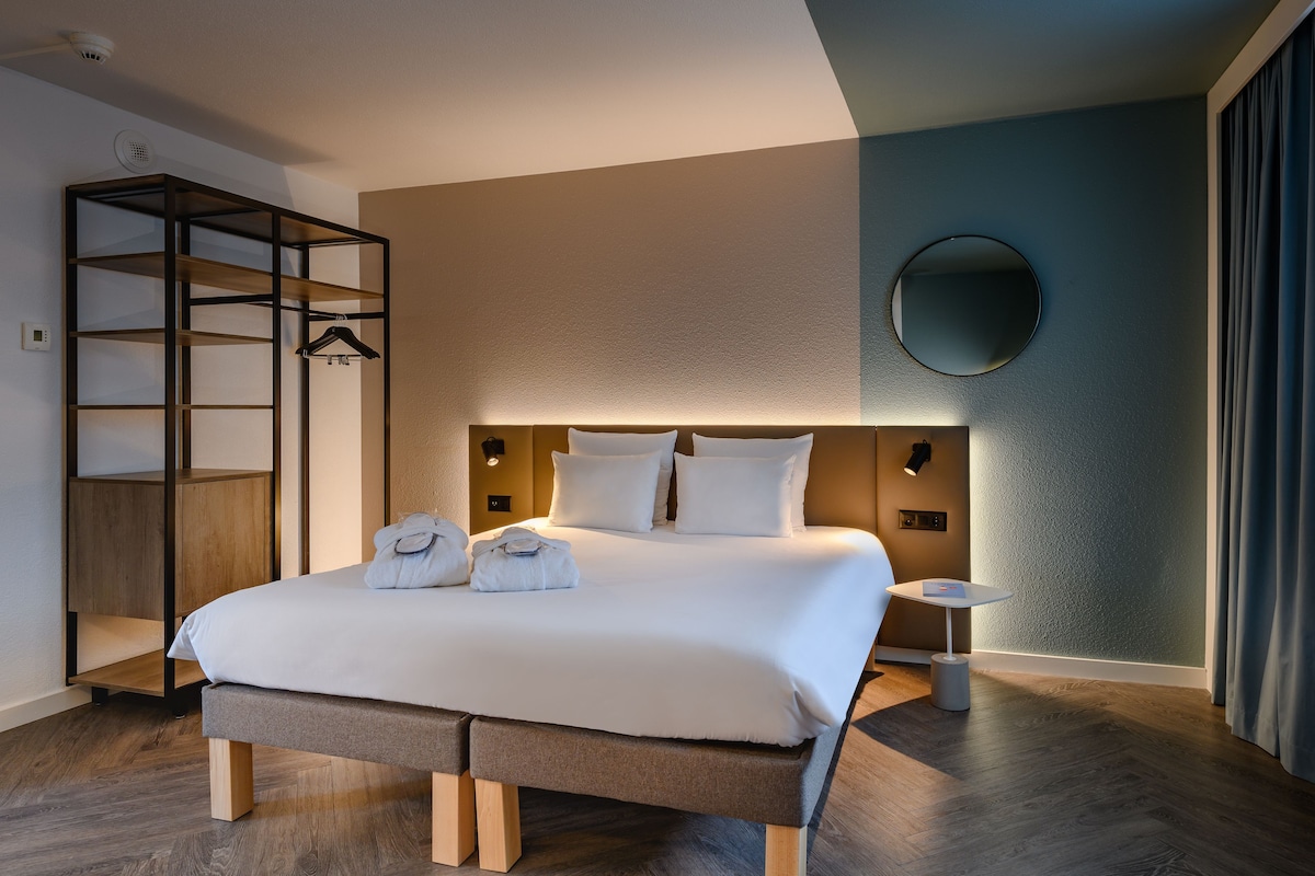 Siististi järjestetty hotellihuone, jossa on suuri sänky, yöpöydät, vaatekaappi ja pyöreä peili seinällä.