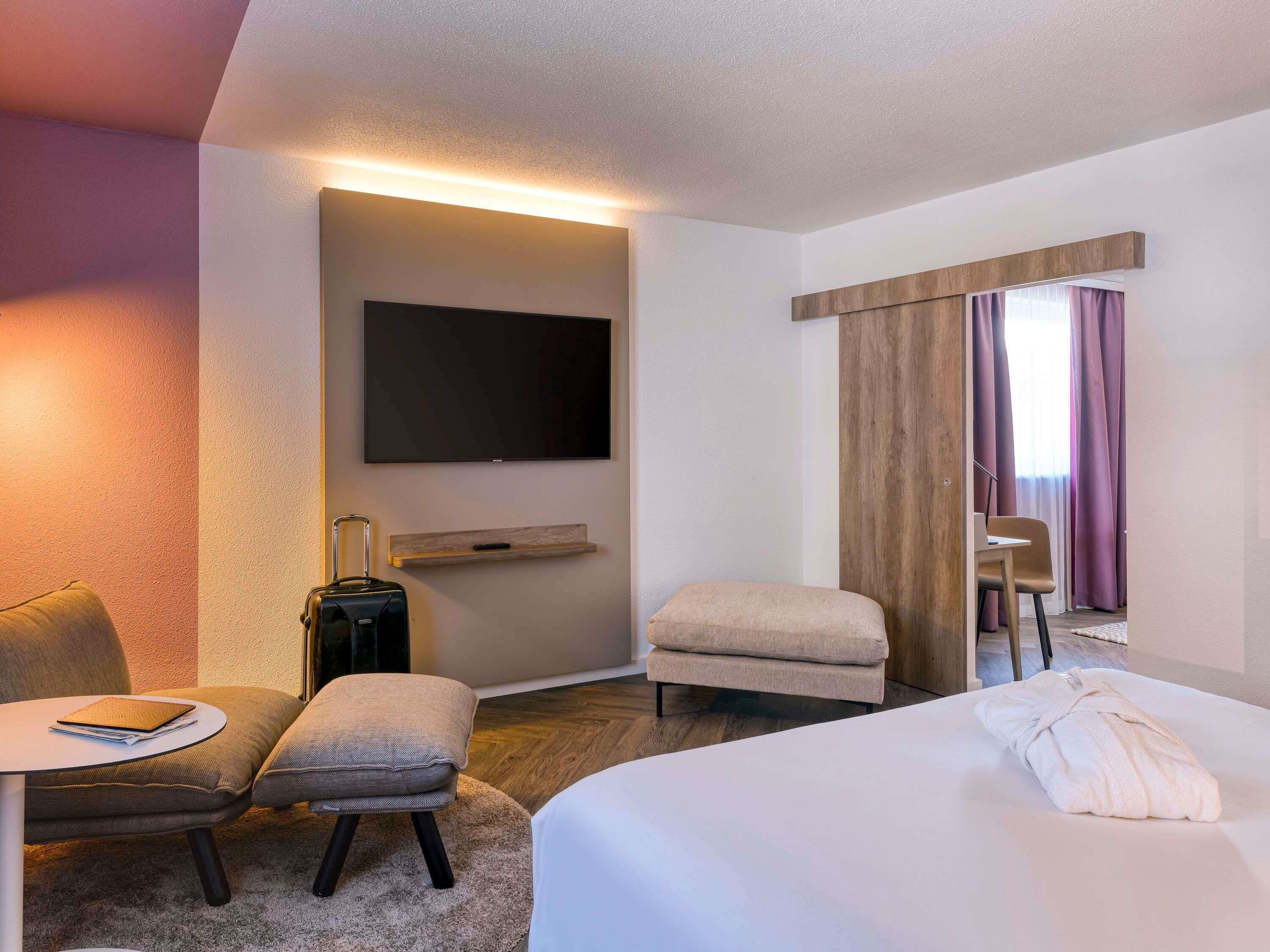 A hotel room with a bed, a desk with a chair, a small table, a television, and a window with curtains.