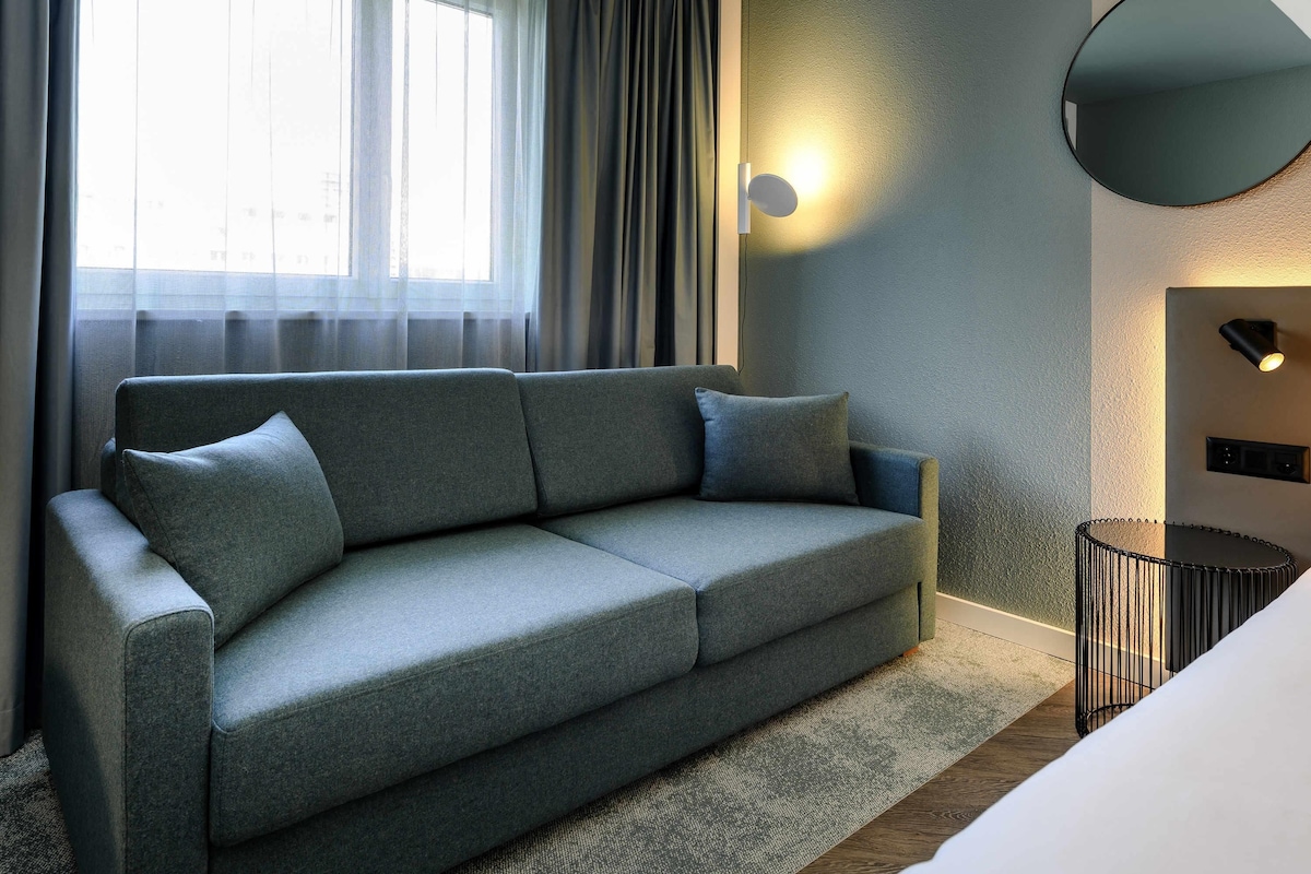 Une chambre d’hôtel moderne avec un canapé gris, un miroir rond et une petite table d’appoint.