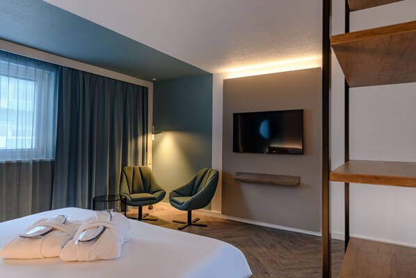 Una habitación de hotel con una cama, dos sillones, un televisor en la pared y un estante de madera.