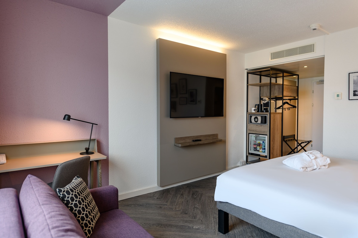 Moderni hotellihuone, jossa on sänky, sohva, työpöytä lampun kanssa, televisio ja vaatekaappi.
