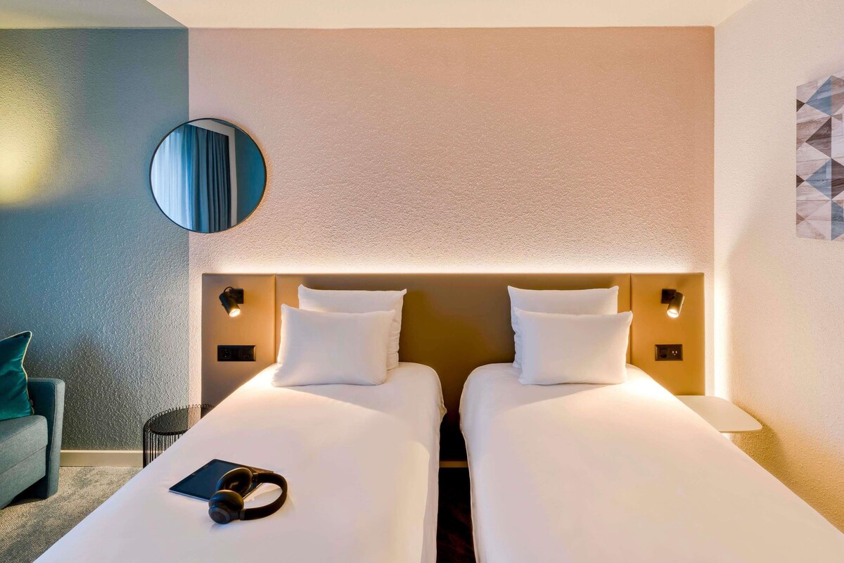 Une chambre d’hôtel avec deux lits, un miroir rond, des lampes de chevet, une petite table et une chaise.