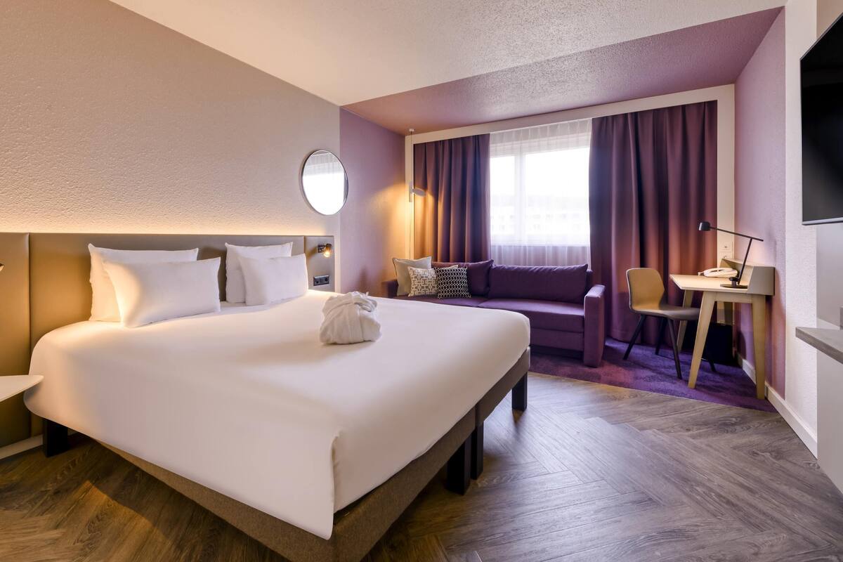 Hotellihuone, jossa on suuri sänky, työpöytä tuolilla, pyöreä peili ja ikkuna, jossa on verhot.