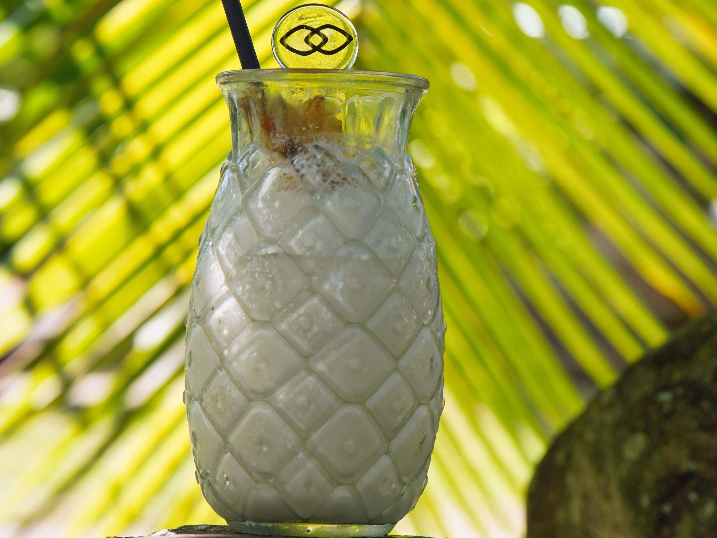 A glass with a textured pattern, containing a beverage, with a straw and a lid with a logo, set against a palm leaf background.