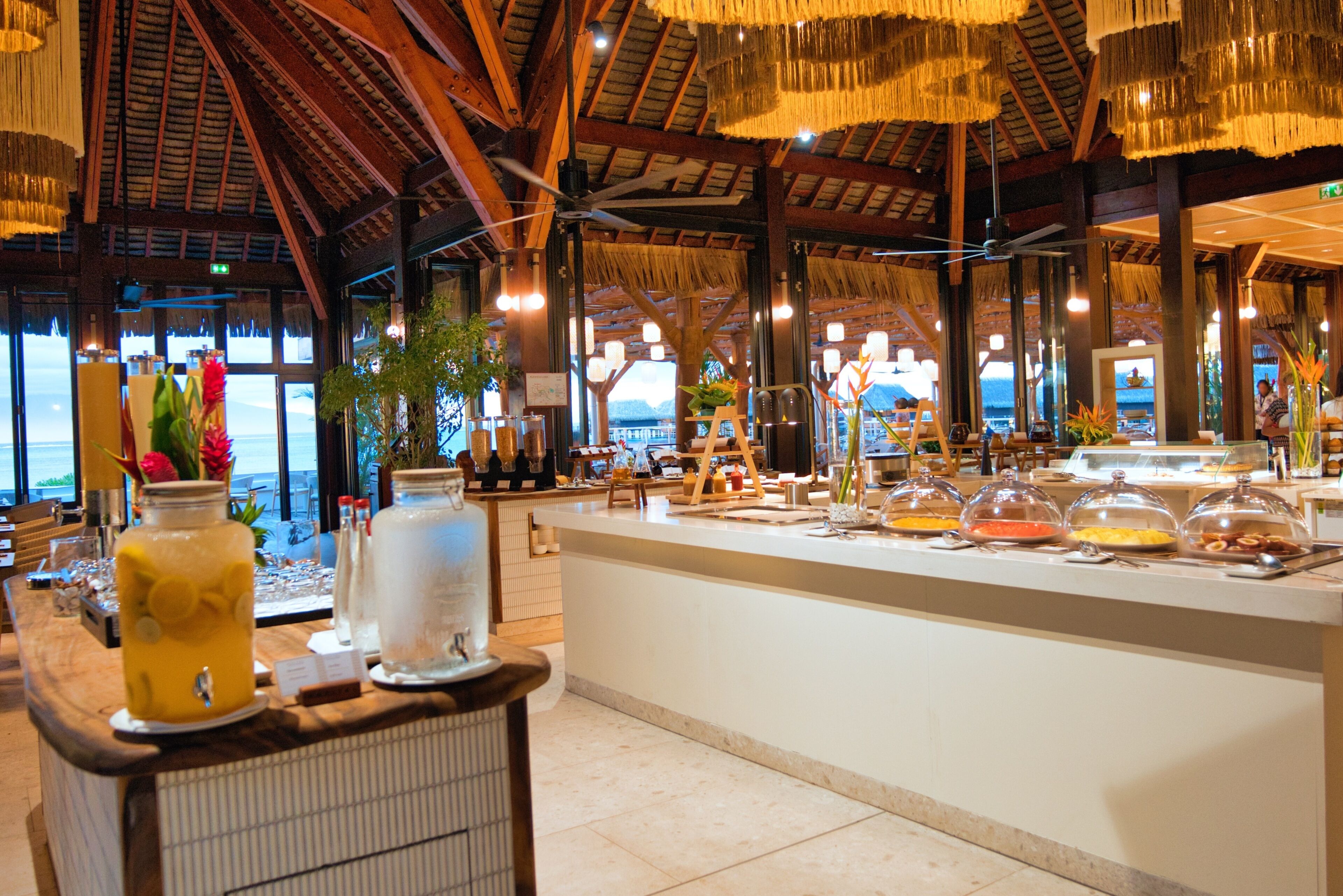 A buffet area with a variety of food items under glass domes, a bar area with a drink dispenser, and a view of the beach through large windows.