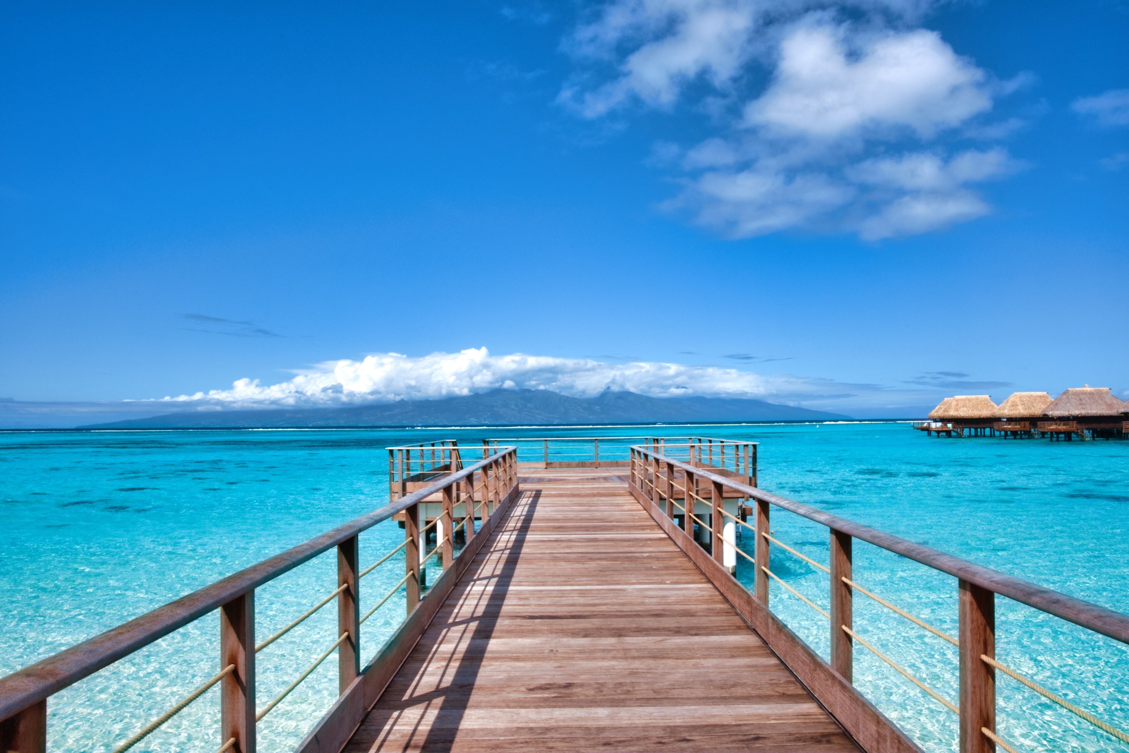 Un muelle de madera que conduce a cabañas sobre el agua, con agua turquesa cristalina y una montaña al fondo.