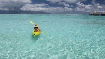 Una persona practica kayak en aguas turquesas cristalinas, con bungalós sobre el agua al fondo.