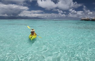 Una persona practica kayak en aguas turquesas cristalinas, con bungalós sobre el agua al fondo.