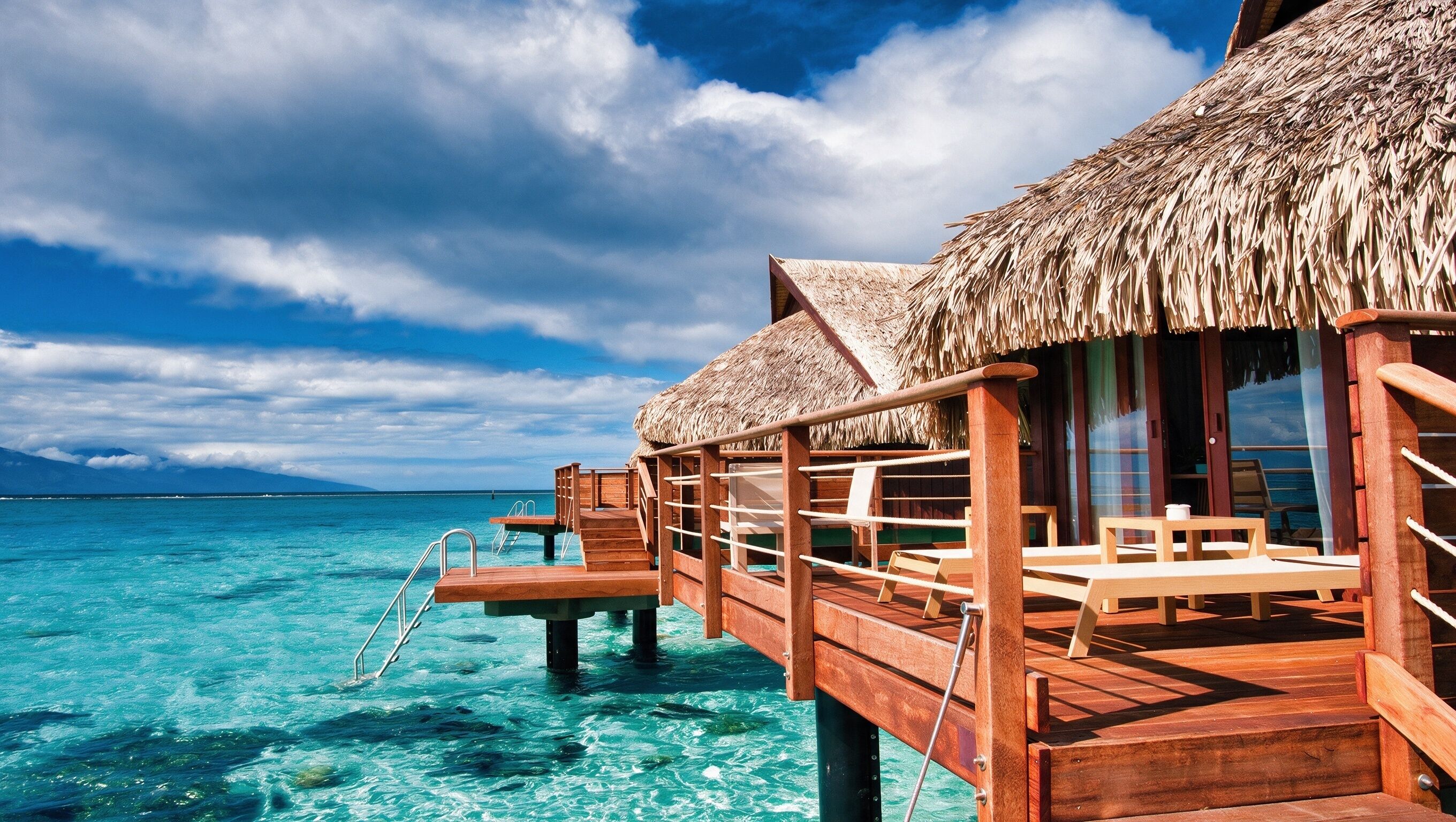 A wooden overwater bungalow with a thatched roof, wooden deck, and a clear view of the turquoise sea.