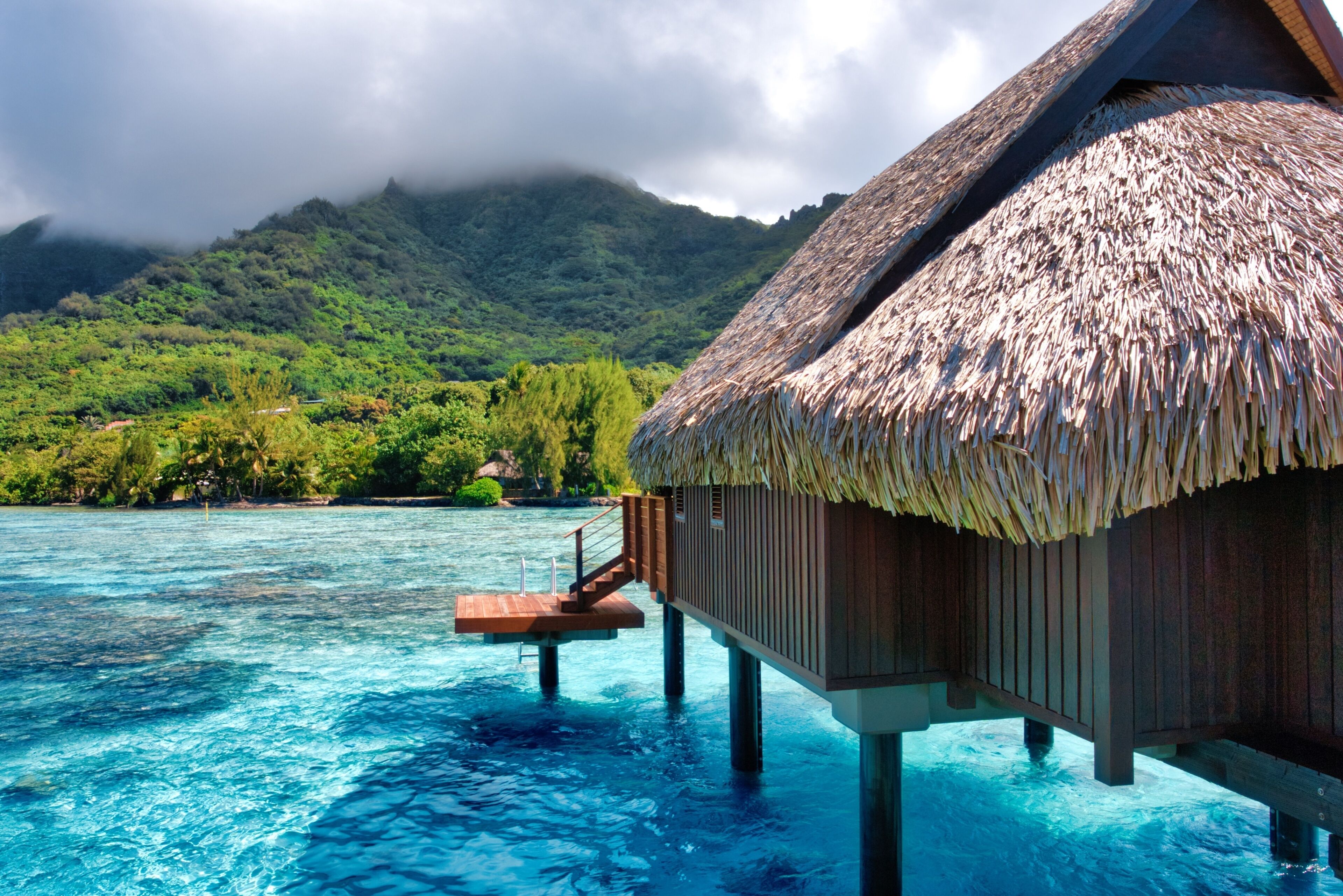 An overwater bungalow with a thatched roof, wooden deck, and a view of the turquoise sea and lush green hills.