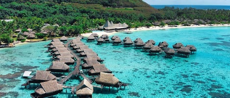 An overwater bungalows area with thatched roofs, clear turquoise water, and a lush green backdrop.