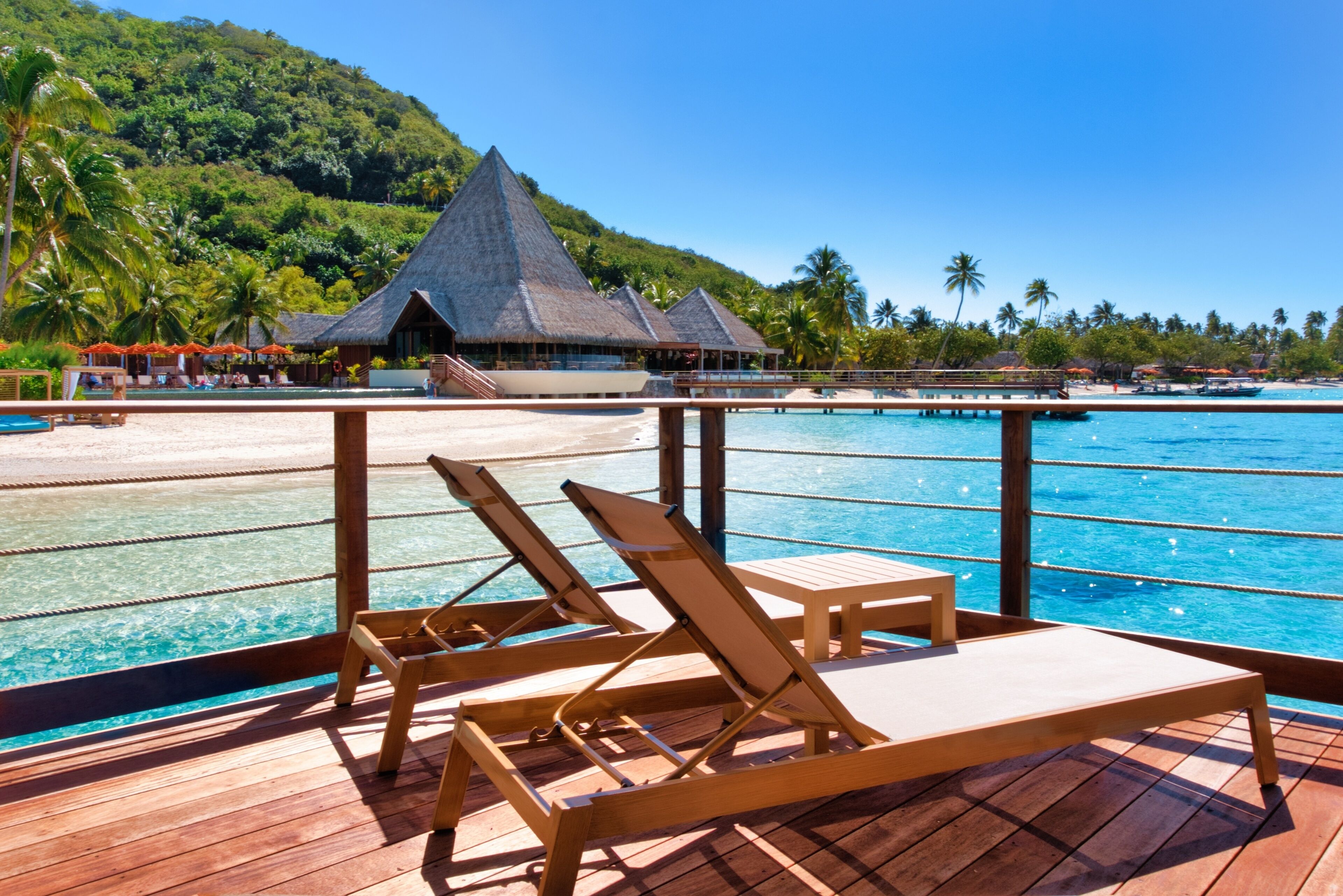 A wooden deck with two lounge chairs overlooking a clear blue sea and a tropical beach with thatched-roof buildings.