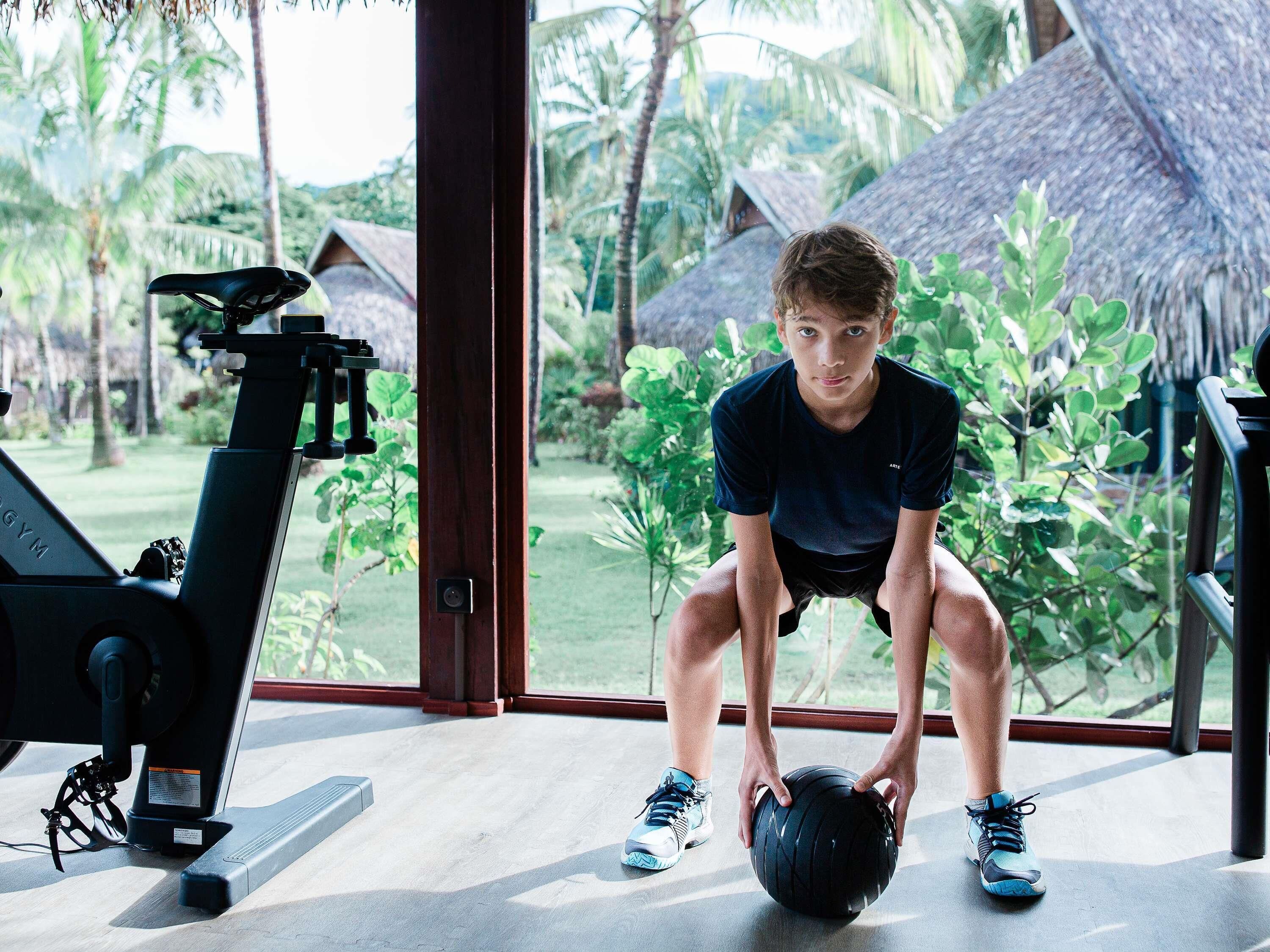 A person performing a squat exercise with a medicine ball in a room with a treadmill and large windows overlooking a garden.