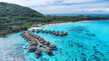 Vue aérienne d’un complexe hôtelier avec des bungalows sur pilotis reliés par des passerelles, entouré d’eaux turquoise cristallines et d’un paysage verdoyant luxuriant.