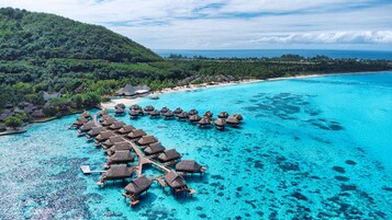 An aerial view of a resort with overwater bungalows connected by walkways, surrounded by clear turquoise waters and a lush green landscape.