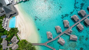 Vista aérea de un resort con bungalows sobre el agua conectados por una pasarela de madera, una piscina y una playa.