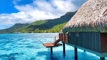 An overwater bungalow with a thatched roof, wooden deck, and a view of the ocean and a mountain in the background.