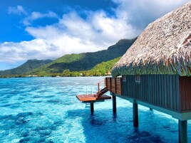 An overwater bungalow with a thatched roof, wooden deck, and a view of the ocean and a mountain in the background.
