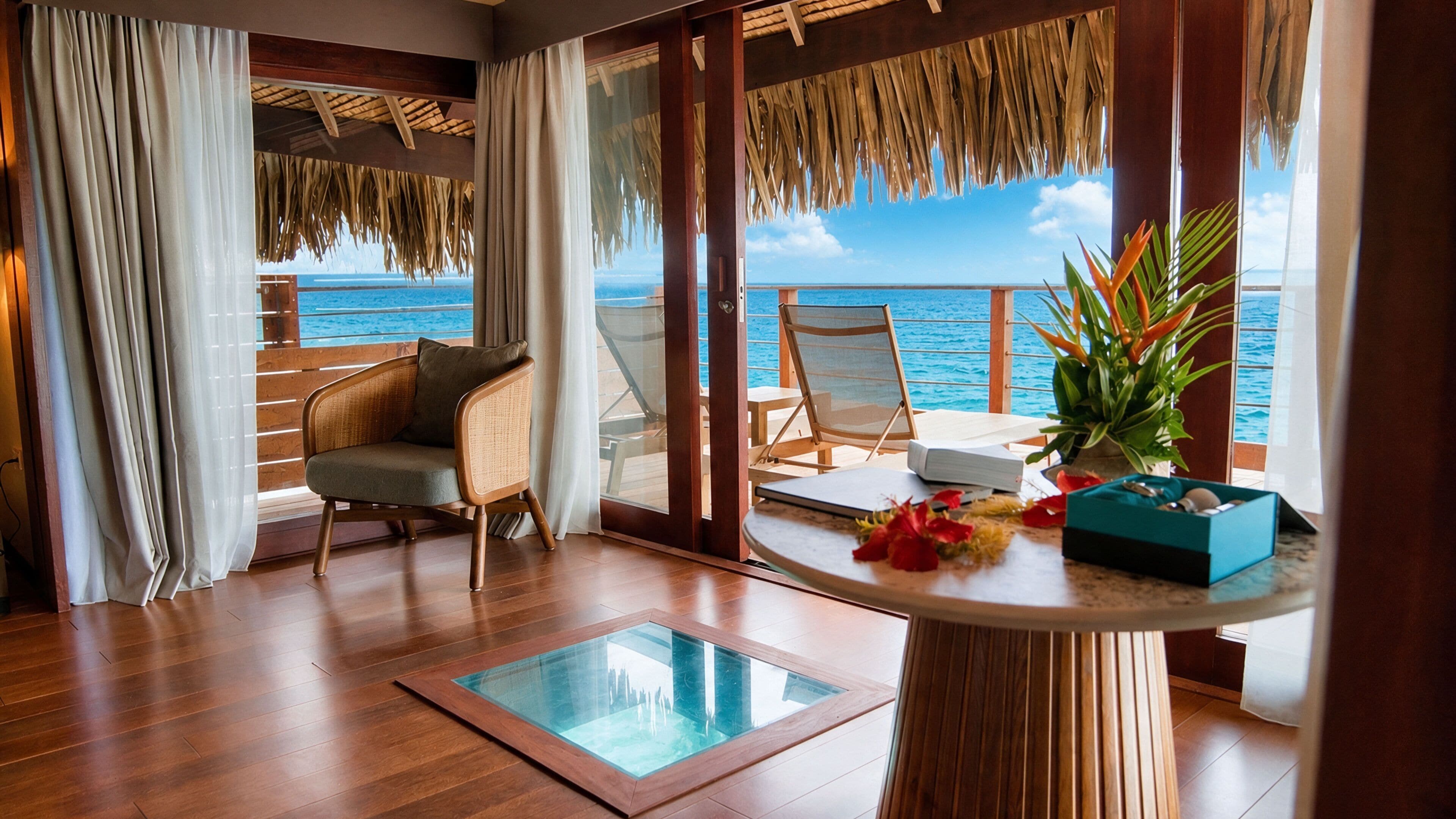 A room with a wooden floor, a glass table, a chair, a small table with a plant, and a view of the ocean.