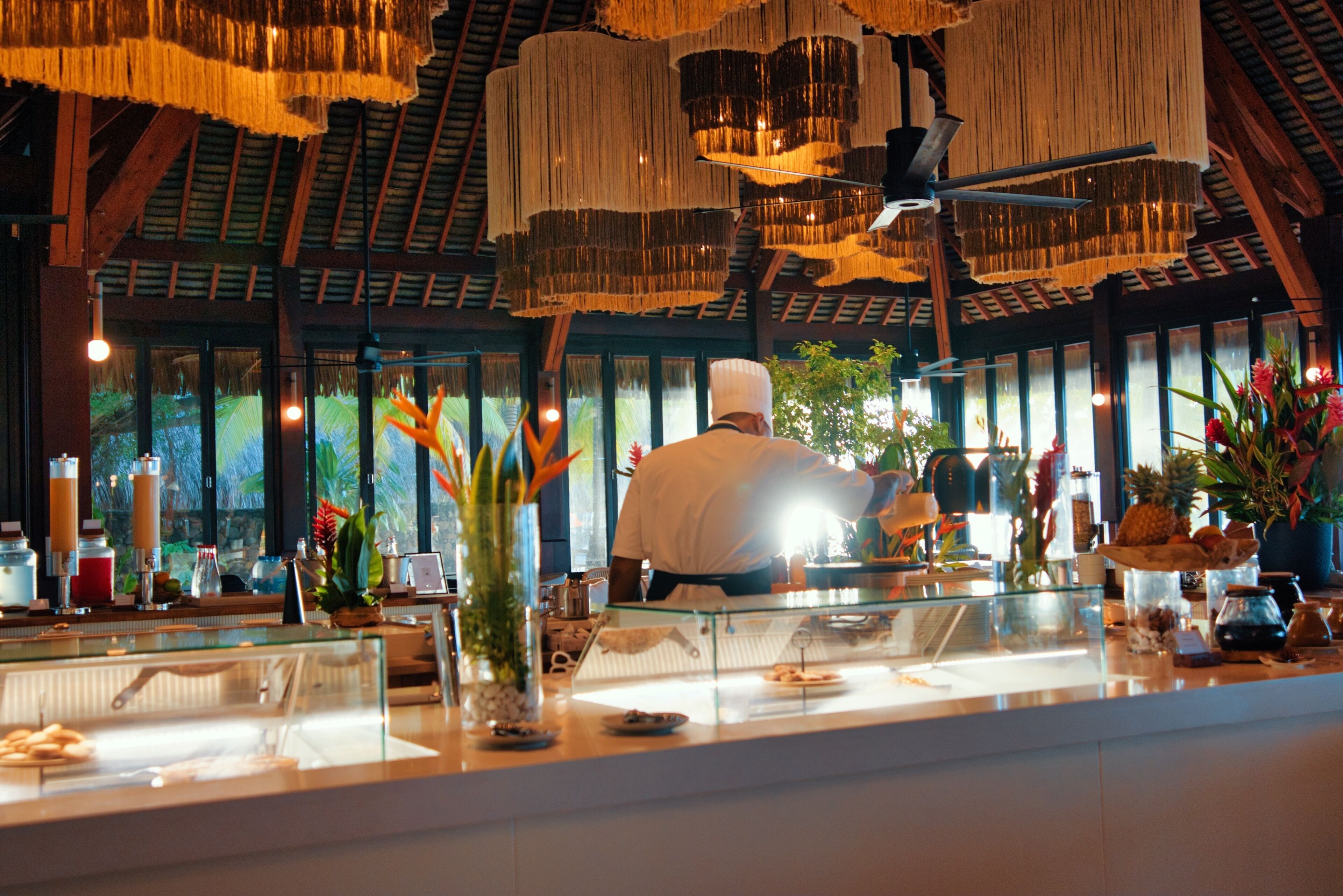 Un chef preparando comida en un restaurante de temática tropical, con techo de paja y ventanas amplias.