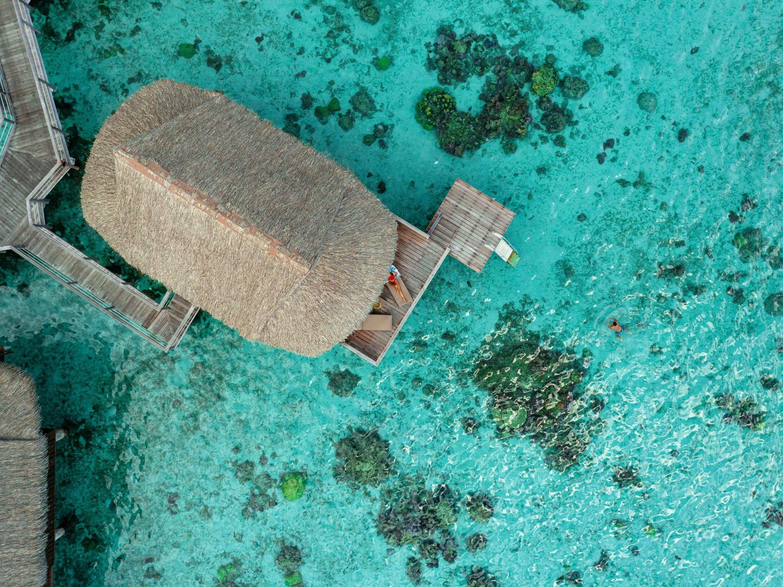 An aerial view of a resort with a thatched-roof structure connected to a wooden deck over turquoise waters.
