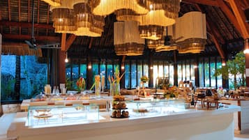Amplia habitación con techo alto y candelabros colgantes, un mostrador de buffet con diversas opciones de comida, y un gran ventanal con vistas al exterior.