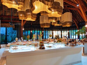 Amplia habitación con techo alto y candelabros colgantes, un mostrador de buffet con diversas opciones de comida, y un gran ventanal con vistas al exterior.
