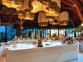 A spacious room with a high ceiling and hanging chandeliers, a buffet counter with various food items, and a large window providing a view of the outdoors.