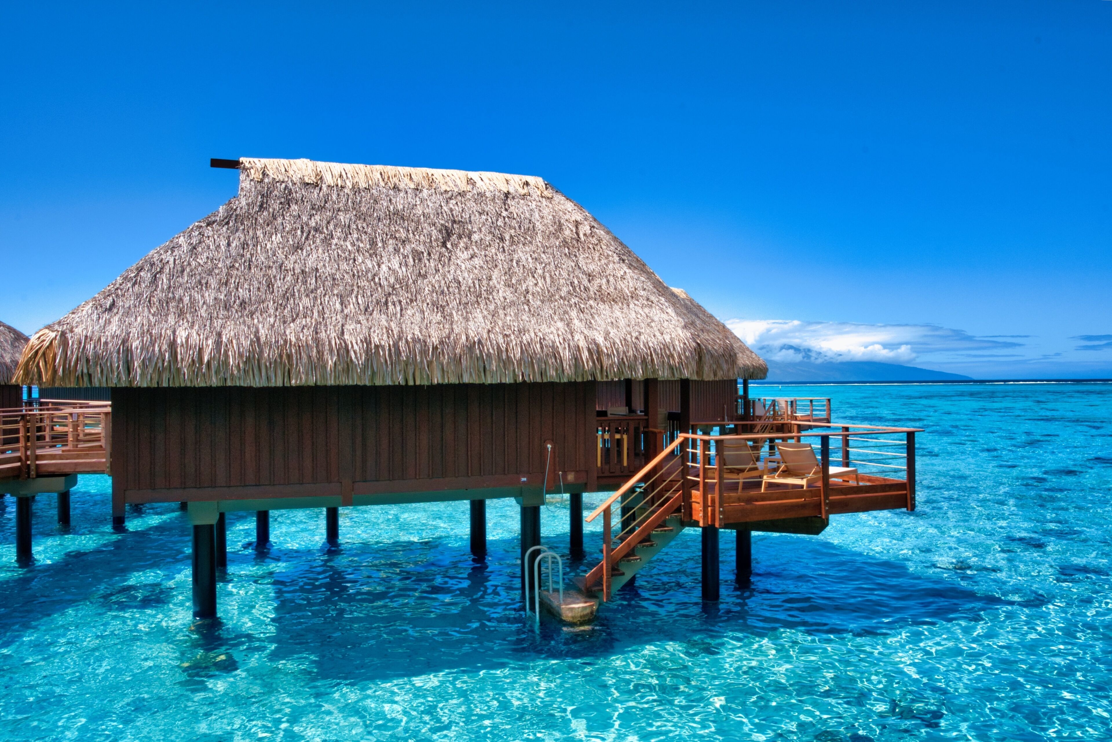 An overwater bungalow with a thatched roof, wooden deck, and clear blue water.
