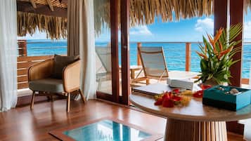 A room with a wooden floor, a glass table, a chair, a small table with a plant, and a view of the ocean.