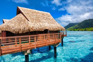 A thatched-roof overwater bungalow with a wooden deck extending into clear blue water.