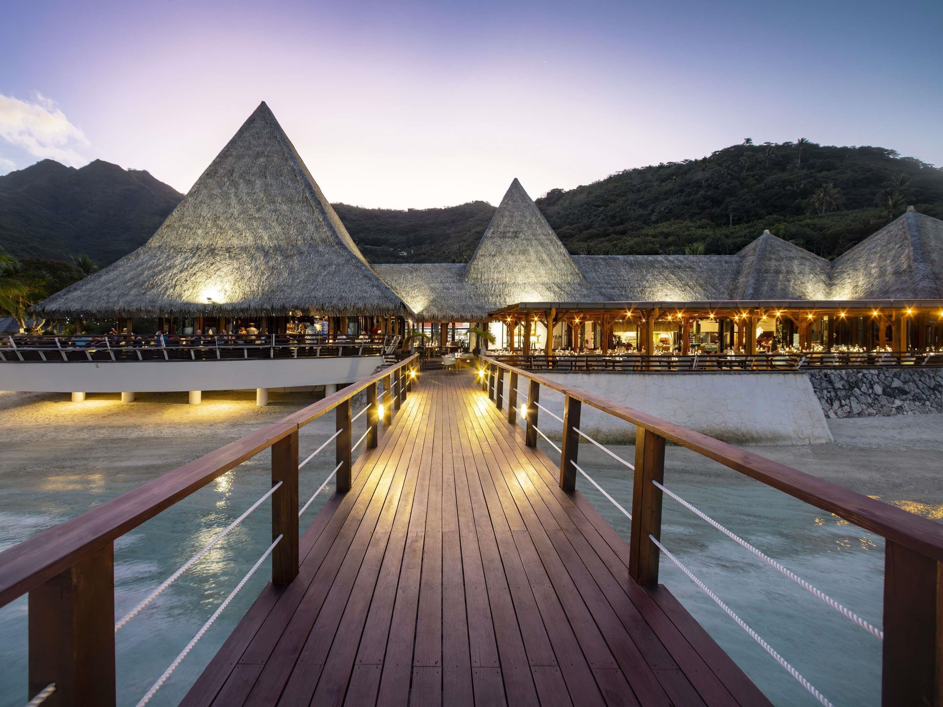 A wooden deck leading to thatched-roof structures with outdoor seating, overlooking a body of water and a mountainous backdrop.