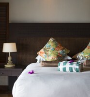 Una cama bien hecha con almohadas estampadas con flores, una toalla a cuadros y una flor morada sobre la cama.