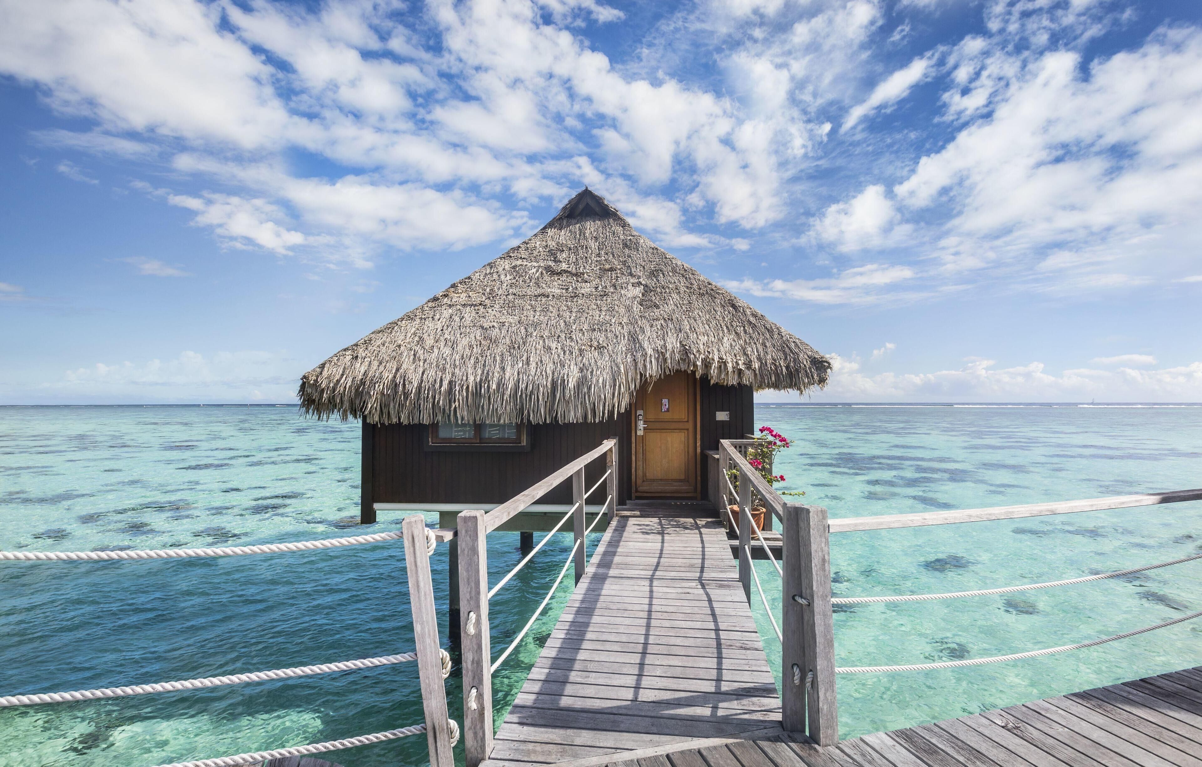 A wooden overwater bungalow with a thatched roof, a wooden deck, and clear turquoise water.