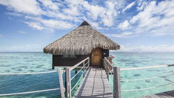 A wooden overwater bungalow with a thatched roof, a wooden deck, and clear turquoise water.